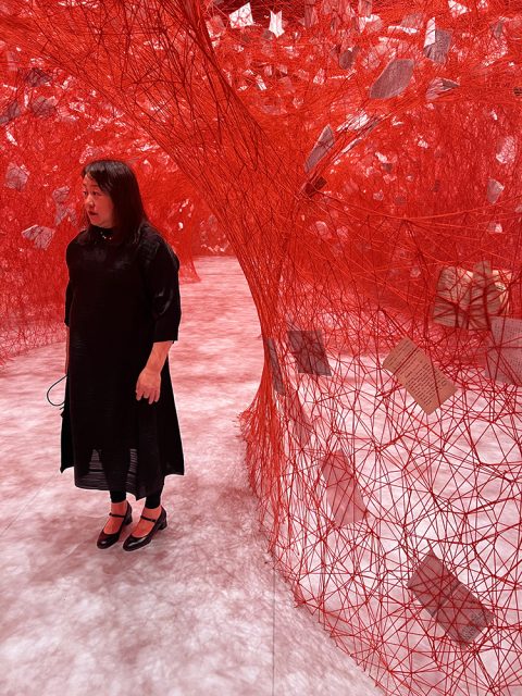 A woman stands near an intricate installation of red thread and suspended papers woven between tree-like structures in a gallery space.