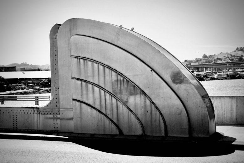 Large metallic structure with curved, ribbed panels sits on a bridge, with a river and distant buildings visible in the background under bright sunlight.