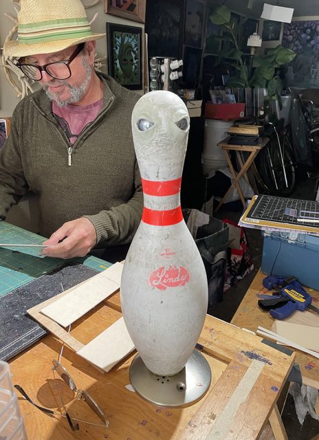 A man wearing glasses and a hat works at a cluttered table. In the foreground, a bowling pin with painted eyes is mounted on a stand.
