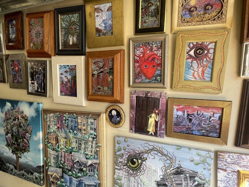 A wall covered with various framed paintings, mostly surreal and colorful, including images of eyes, faces, houses, and abstract patterns.