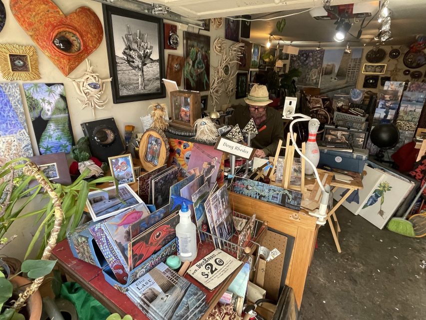 A cluttered art studio filled with colorful paintings, eclectic decor, books, and a man wearing a hat sitting at a desk surrounded by creative materials.