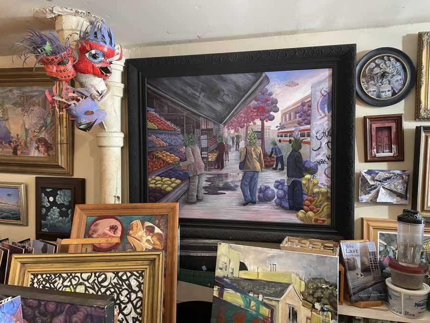 Art studio with various framed paintings, including a large market scene, colorful masks on the wall, and art supplies on a cluttered table.