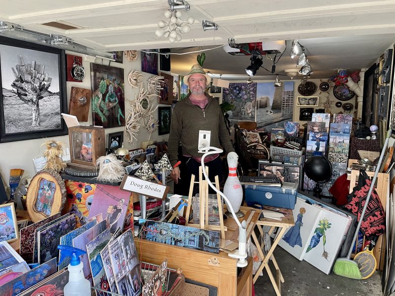 A man stands in a cluttered room filled with various artworks, sculptures, and miscellaneous objects, with paintings covering the walls and tables crowded with art pieces.