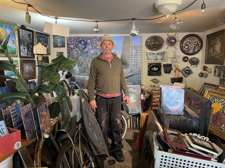 A man in a hat stands in a cluttered room filled with paintings, art supplies, and various art pieces covering the walls and floor.