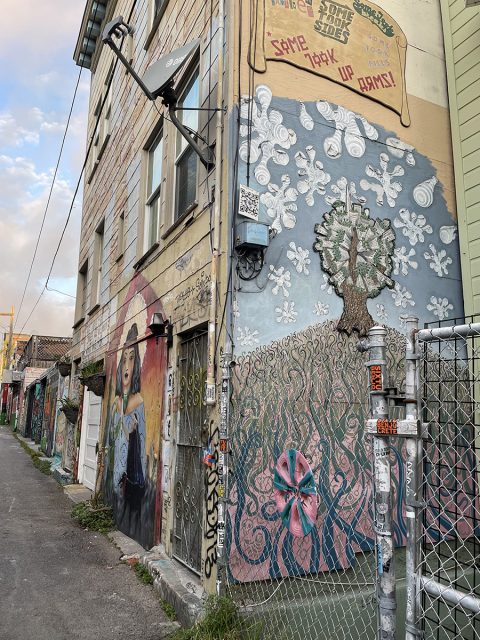 A building in an alley features colorful murals, including a woman in blue, a tree with white shapes, and pink flowers. A metal fence is in the foreground.