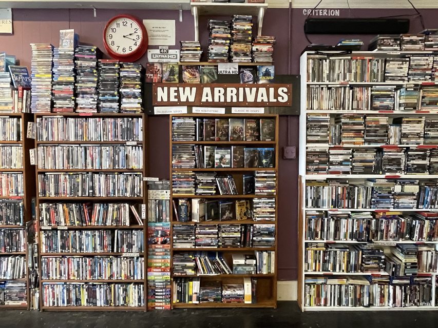 Bookshelves filled with CDs and DVDs, with a central "New Arrivals" sign and a wall clock above the shelves on the left.