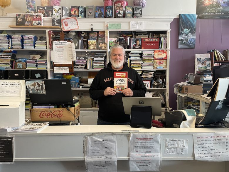 A man stands behind a bookstore counter holding a copy of "Used and Rare." Shelves of books and various items fill the background.