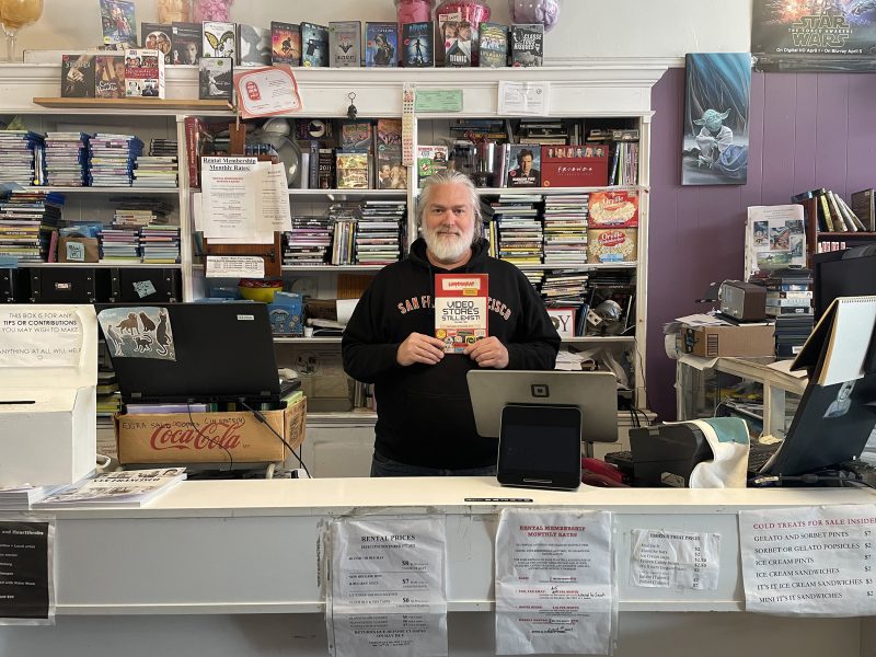A man stands behind a bookstore counter holding a copy of "Used and Rare." Shelves of books and various items fill the background.
