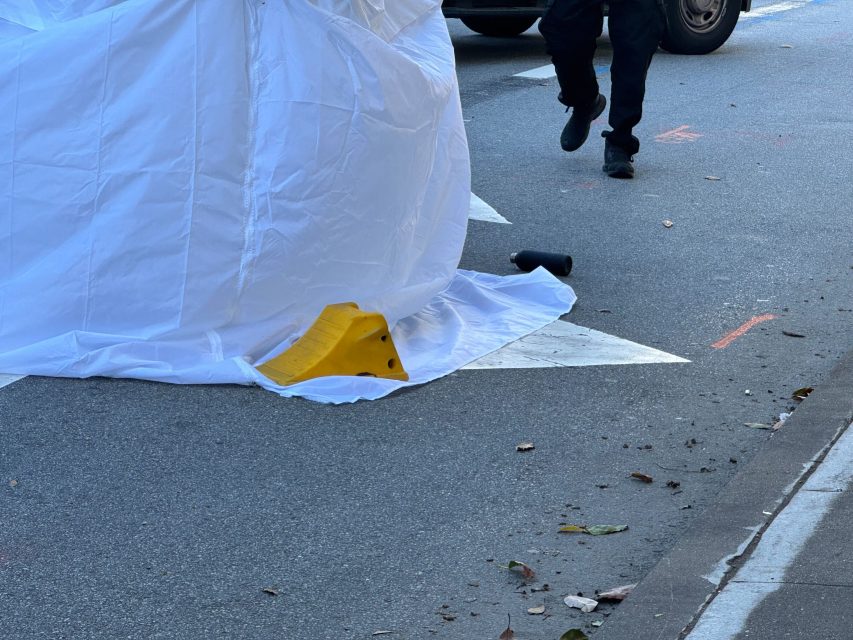 A white tarp covers an object on a street next to a yellow barrier, while a person in dark clothing stands nearby. Some debris and leaves are scattered on the road.