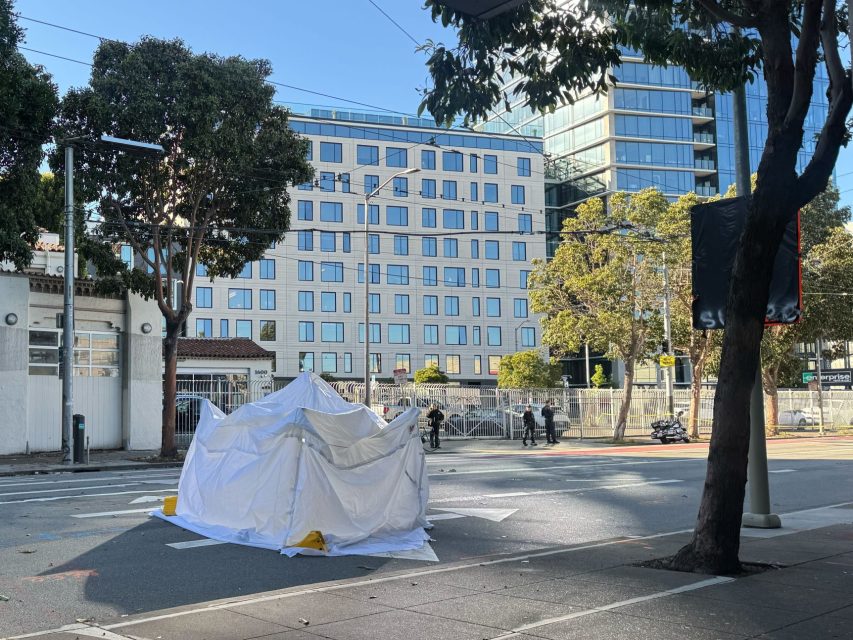 A white tent structure covers an area on a city street, with trees, a sidewalk, and a modern building in the background.