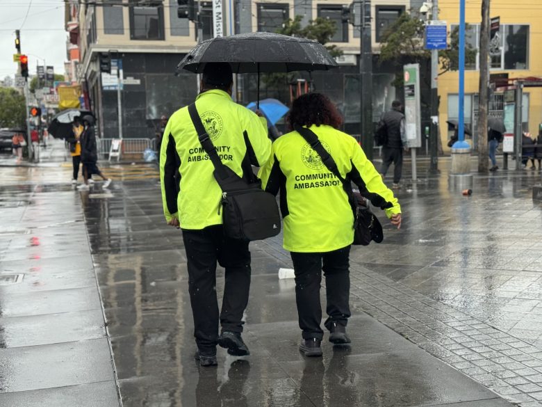 Two people wearing bright yellow "Community Ambassador" jackets walk together under an umbrella on a rainy city street.