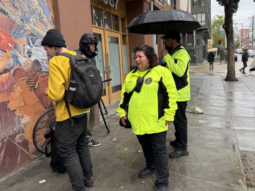 Three people in bright yellow jackets stand on a wet sidewalk talking to two individuals near a mural; one holds an umbrella, and a bicycle is visible.