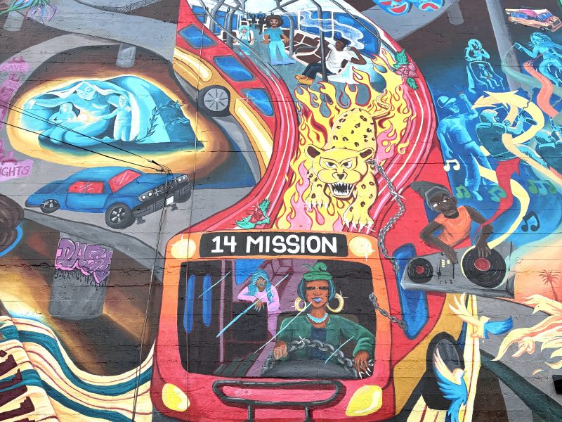 Colorful mural featuring a red city bus labeled "14 Mission," vibrant urban scenes, a tiger, city skyline, people, and surreal elements blending together on a large wall.