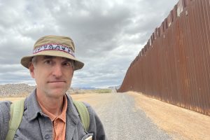 A man wearing a hat and backpack stands on a gravel road next to a tall metal border fence under a cloudy sky.