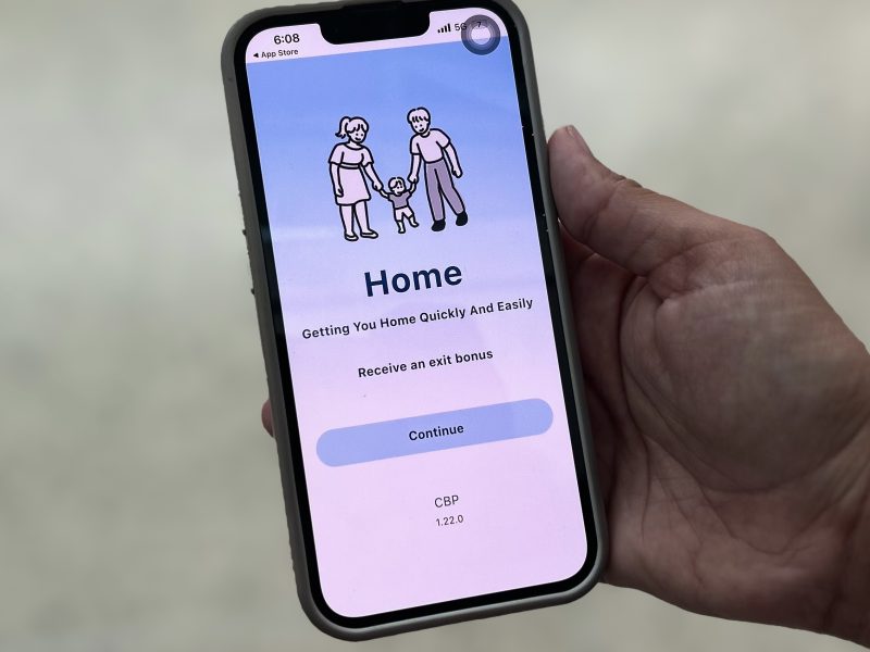 A hand holding a smartphone displaying an app screen with a cartoon family, the word "Home," and a "Continue" button—designed to support every asylum-seeker on their journey to safety.