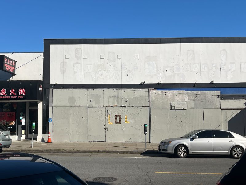 A gray, weathered building facade with faded lettering and boarded-up windows stands beside a restaurant entrance; cars are parked along the street.