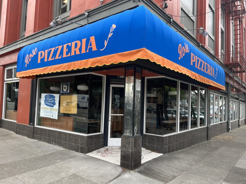 A corner pizzeria in Richmond with a blue and orange awning reading "Pizzeria" and "Rose's," glass windows, and a sidewalk in front.
