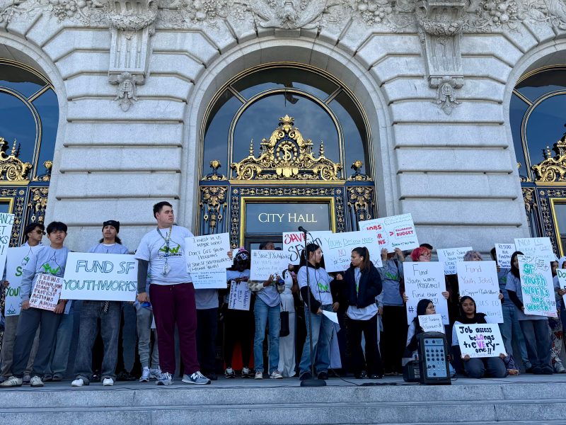 A group of people stand on the steps of City Hall holding signs advocating for youth programs and funding.