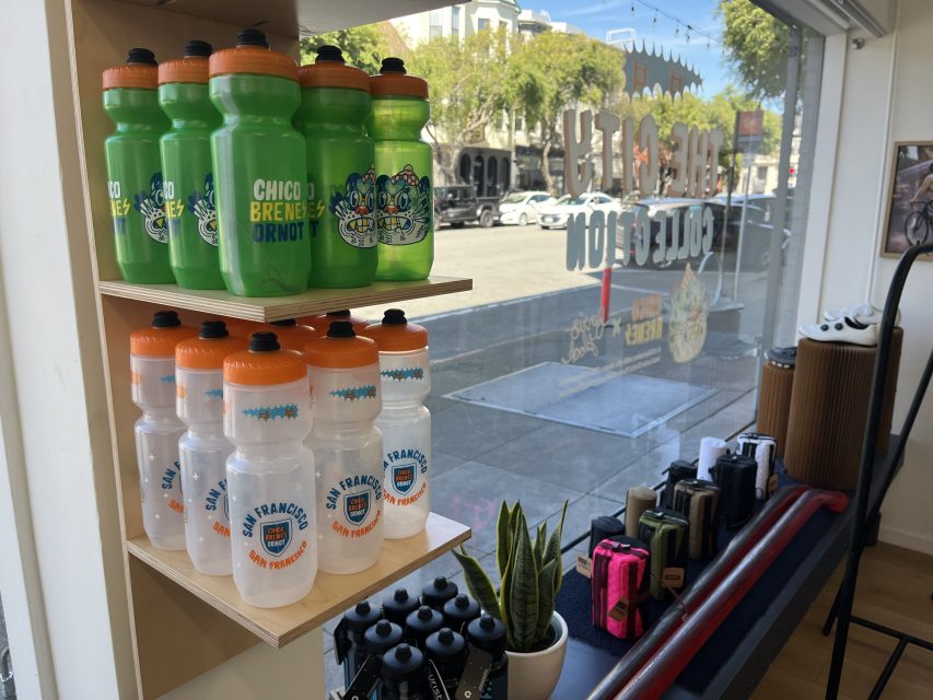 Shelves display green and clear water bottles with city logos, small plants, and colorful handlebar bags in a bike shop with large windows facing the street.