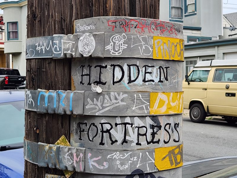 A wooden utility pole wrapped with metal bands at intervals, each covered in graffiti and stickers, with the words "HIDDEN FORTRESS" painted in black.