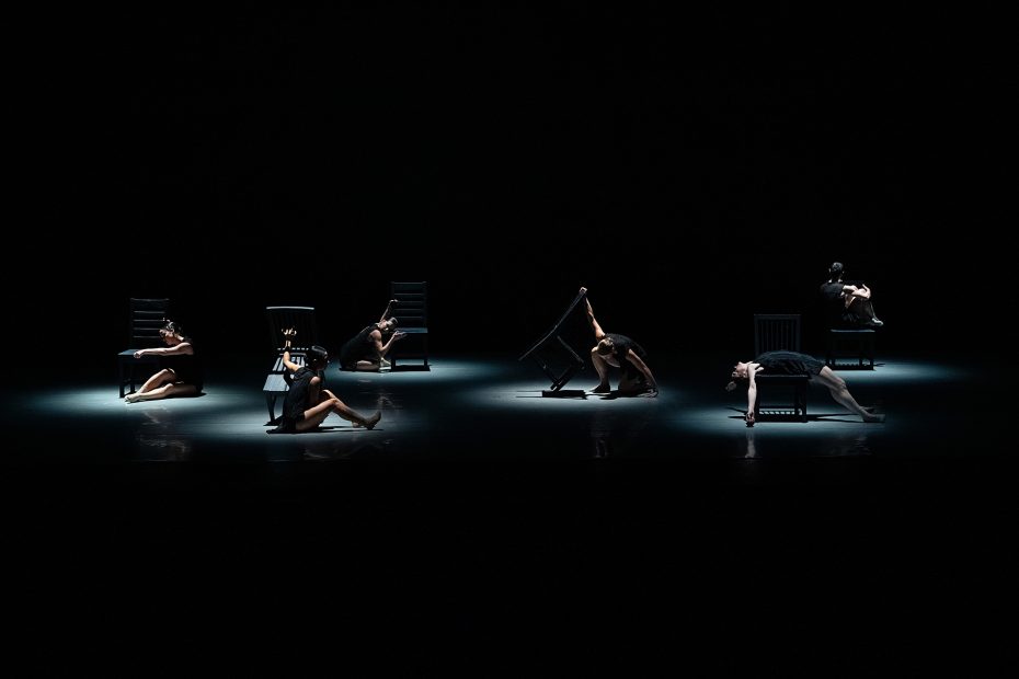 Six dancers perform on a dimly lit stage, each interacting with a chair in varied poses under focused spotlights against a dark background.