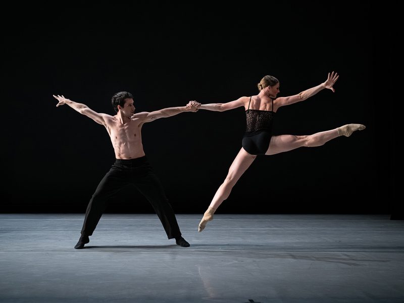 Two ballet dancers perform on stage; the male dancer stands with arms outstretched while holding hands with the female dancer, who is leaping gracefully with legs extended.