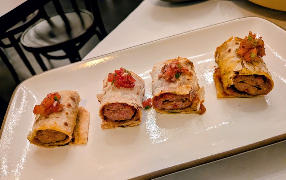 Four rolled tortillas filled with sausage and topped with a small amount of salsa, served on a rectangular white plate.