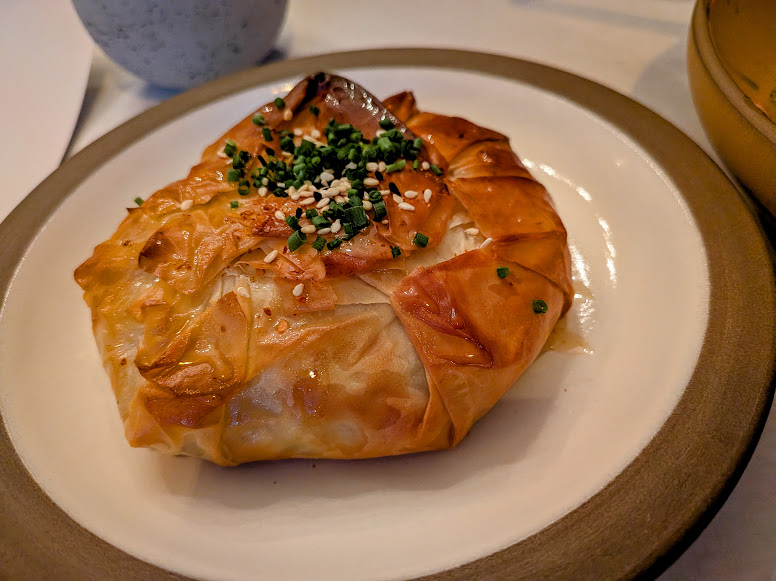 A golden-brown baked pastry topped with chopped chives and sesame seeds is served on a white plate with a brown rim.