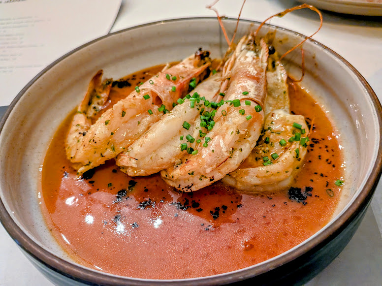 A bowl of grilled shrimp garnished with chopped chives, served in a reddish-orange sauce.