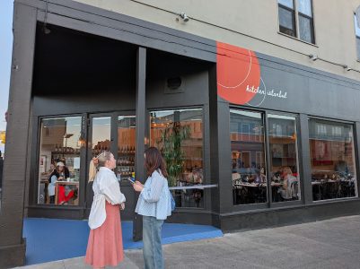 Two women stand talking outside a restaurant with large windows and a red sign that reads "kitchen Istanbul." Diners are visible inside the restaurant.