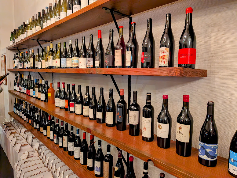 Shelves display rows of assorted wine bottles, with white wines on the top shelf and red wines on the middle and lower shelves, and wrapped bottles lined up along the bottom.