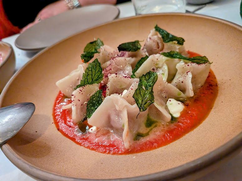 A bowl of dumplings topped with mint leaves, served on a bed of red sauce with white cream dots, placed on a dining table with a spoon nearby.