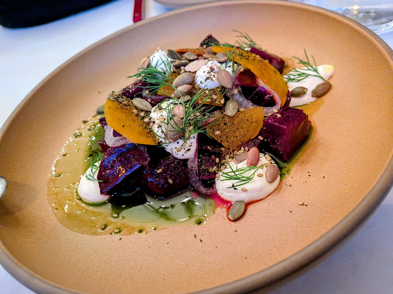 A plated salad with cubed beets, pumpkin seeds, dollops of cream, fresh dill, and thin yellow slices, served on a tan dish with green oil drizzle.