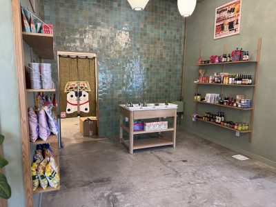 Small specialty grocery store with shelves of packaged goods, a central table displaying items, and a tiled back wall with a decorative curtain over a doorway.