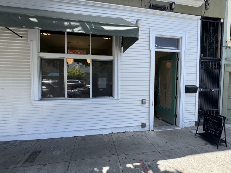 White storefront with a green awning, a large window, an open door, and a black sandwich board sign on the sidewalk.