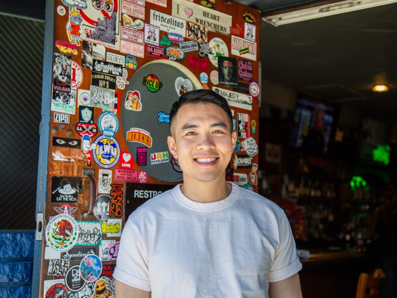 A person in a white t-shirt stands smiling in front of a door covered with colorful stickers at the entrance of a bar or restaurant.