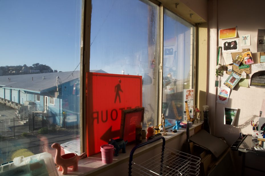 Sunlight streams through a window cluttered with objects and decorations; a red pedestrian detour sign leans against the glass, overlooking buildings and a street outside.
