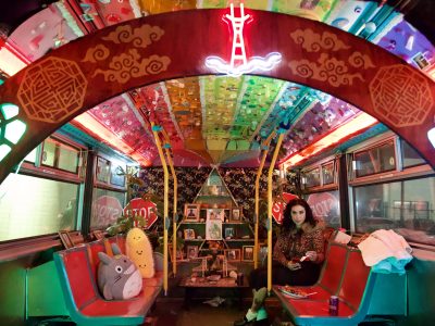 A woman sits in a colorfully decorated bus interior with neon lights, plush toys, and a small altar covered in framed photos and candles at the center.