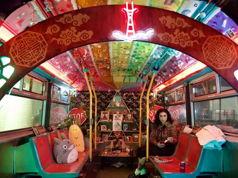 A woman sits in a colorfully decorated bus interior with neon lights, plush toys, and a small altar covered in framed photos and candles at the center.