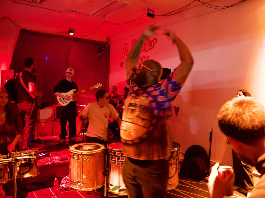 A band performs in a dimly lit room with red lighting as a person in the foreground raises their arms while dancing near drums.