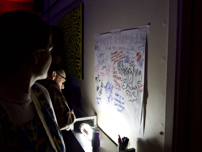 People sign a large birthday card taped to a wall, illuminated by a small light on a ledge beneath it, in a dimly lit room.