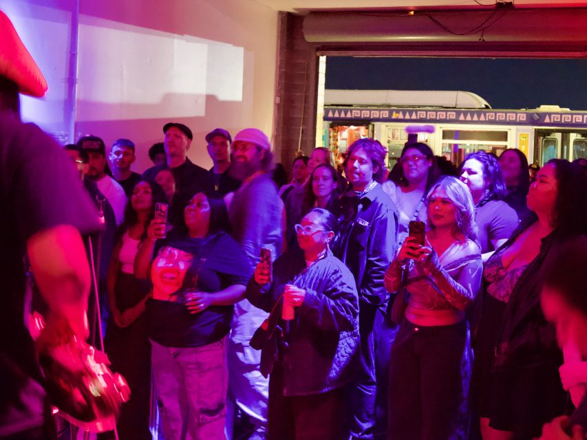 A diverse group of people stand indoors, facing a performer off-frame, many holding up phones. Colorful lighting and a train visible outside suggest an urban setting.