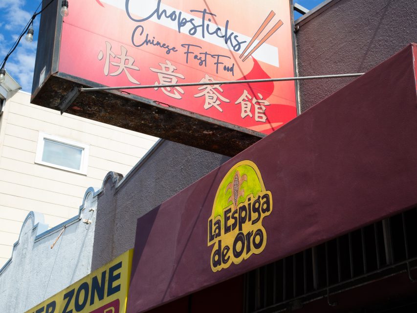 A sign for "Chopsticks Chinese Fast Food" hangs above a maroon awning with the logo "La Espiga de Oro" on a building exterior.
