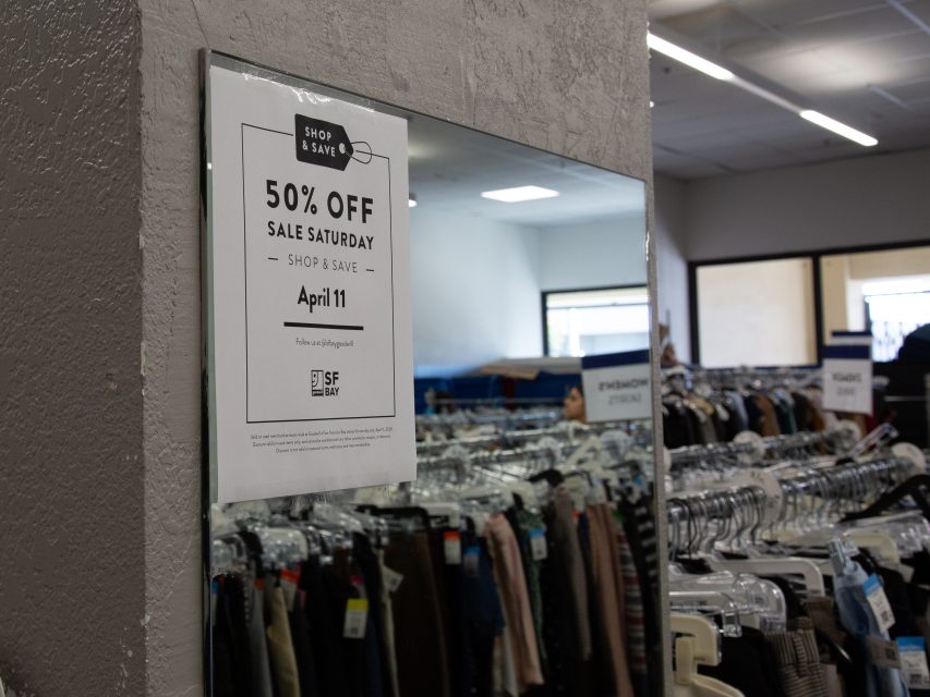 A sign on a mirror in a clothing store announces "50% Off Sale Saturday, April 11." Racks of clothes are visible in the background.