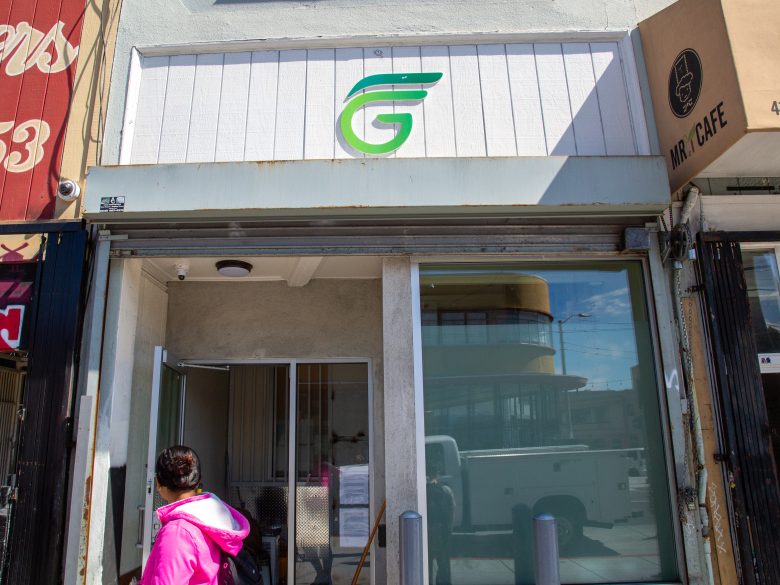 A person in a pink jacket stands outside a storefront with a green and white "G" logo sign above the entrance.