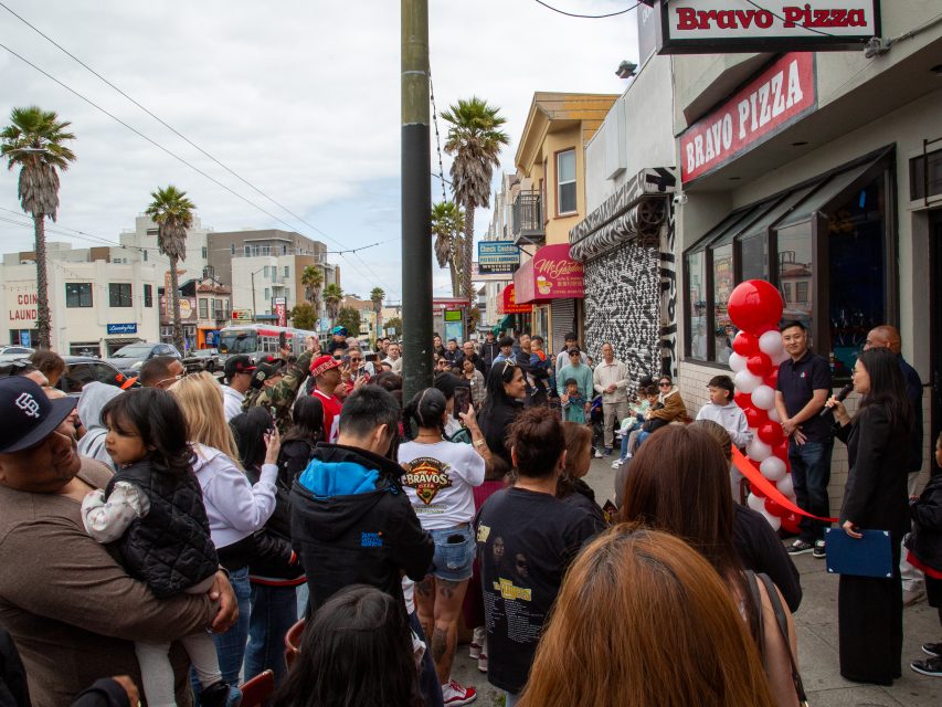 A crowd gathers outside Bravo Pizza on a city street, listening to a person speaking with a microphone; red balloons decorate the entrance, and palm trees line the sidewalk.