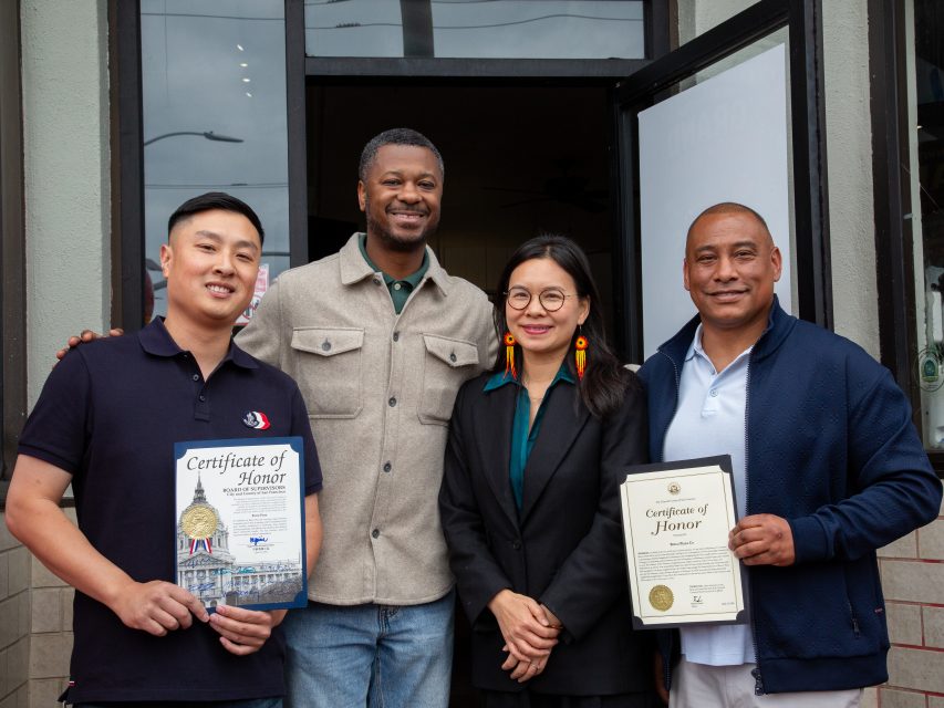 Four people stand together outside a building; two are holding framed certificates of honor and all are facing the camera.