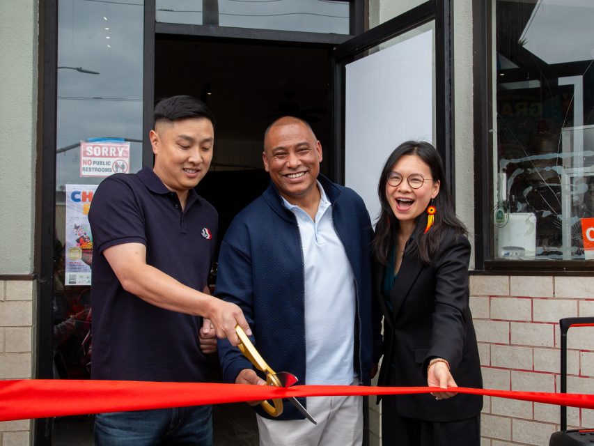 Three people smiling and posing while cutting a red ribbon at the entrance of a building, indicating a ribbon-cutting ceremony or business opening event.