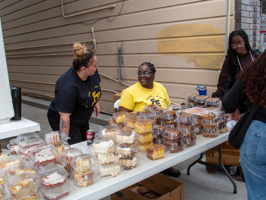 Three people sit and stand behind a table covered with stacks of packaged baked goods at an outdoor event.