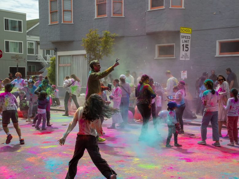 People celebrate outdoors by throwing colored powder in the street; children and adults are covered in color, with buildings and a speed limit sign in the background.